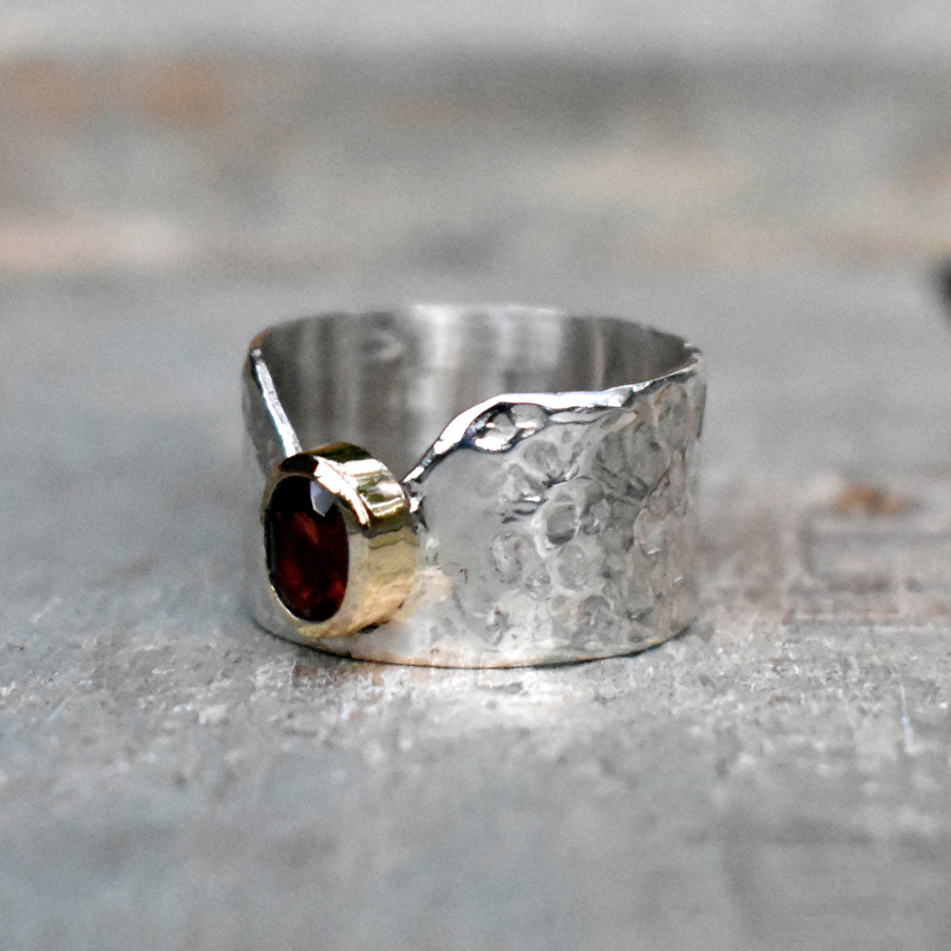 Hammered textured sterling silver band with a V-shape cut-in and a 7mm x 5mm oval Garnet set in a 9ct yellow gold tube. The ring is 12mm  in width.  Please note that all pieces are created on order by Erfdeel Juwele. Allow 2 weeks for manufacture.